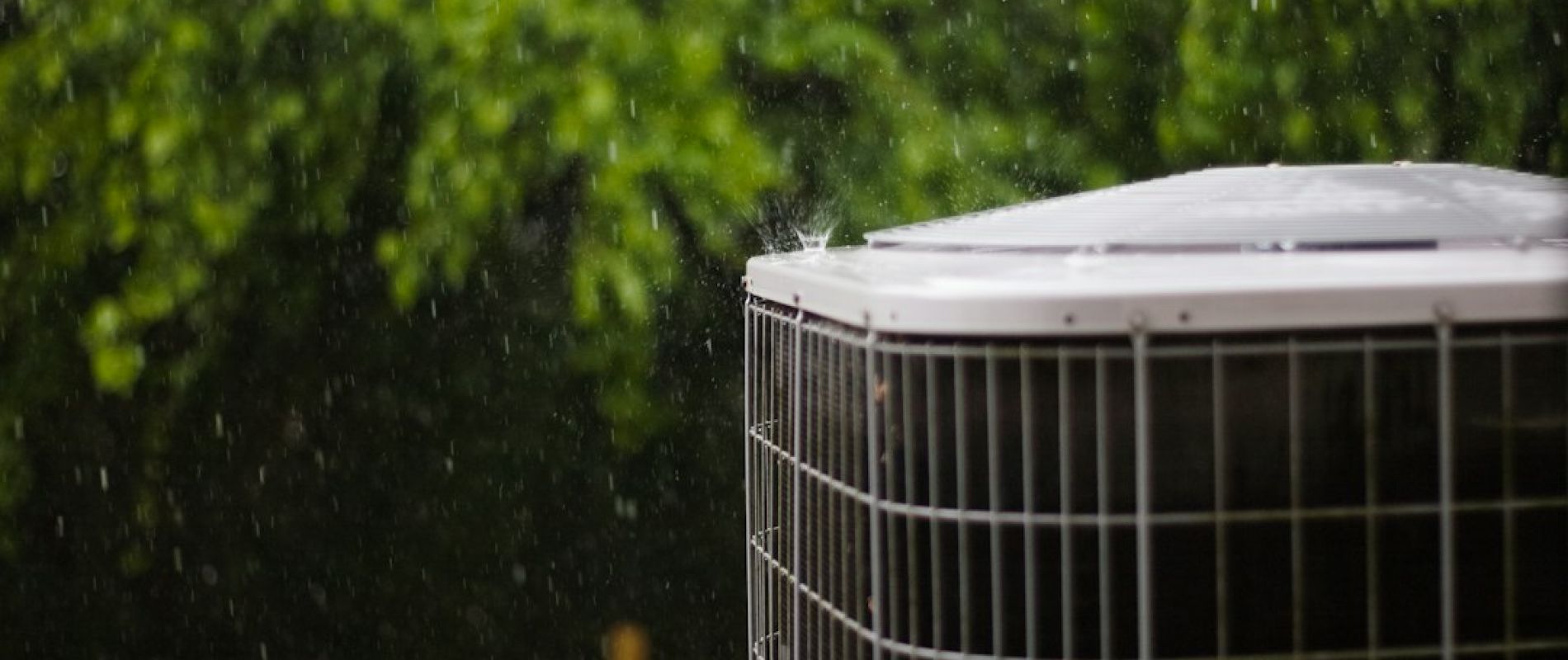 Unidad exterior de aerotermia bajo lluvia con jardín verde de fondo, mostrando la exposición a condiciones climáticas