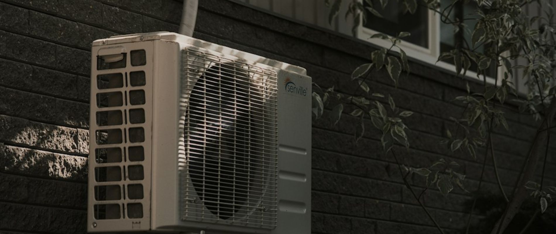 Unidad exterior de bomba de calor aerotérmica instalada en pared junto a tubería y vegetación